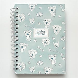 May include: A spiral-bound journal with a light blue cover featuring a repeating pattern of dog illustrations, dog bowls, tennis balls, and chew toys. The text "foster journal" is centered on the cover.