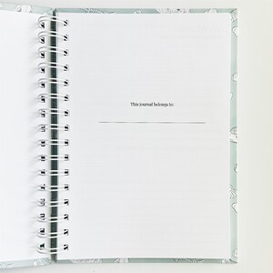 May include: A spiral-bound journal with a light sage green cover featuring a white line-art pattern. The interior page is white with the printed text "This journal belongs to:" followed by a horizontal line for writing a name or information.