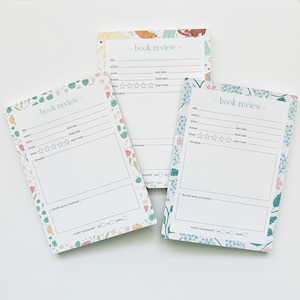 May include: Three floral-themed book review notepads. Each notepad features a white interior with lines for title, author, and rating, and a section for notes. The borders are decorated with colorful flowers and greenery, with the words "- book review -" at the top.
