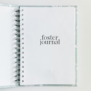 May include: A spiral-bound journal with a light blue cover featuring a white line-art pattern. The interior white page displays the black text foster journal in a serif font. The white metal wire binding runs along the left side of the notebook.
