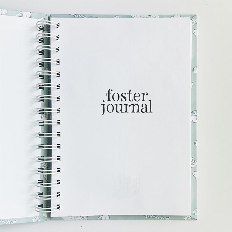May include: A spiral-bound journal with a light blue cover featuring a white line-art pattern. The interior white page displays the black text foster journal in a serif font. The white metal wire binding runs along the left side of the notebook.