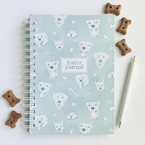 May include: A light blue spiral-bound journal with a repeating pattern of dog faces, tennis balls, and bowls. A central white rectangular label features the text "foster journal" in dark blue. The journal is accompanied by a white pen.