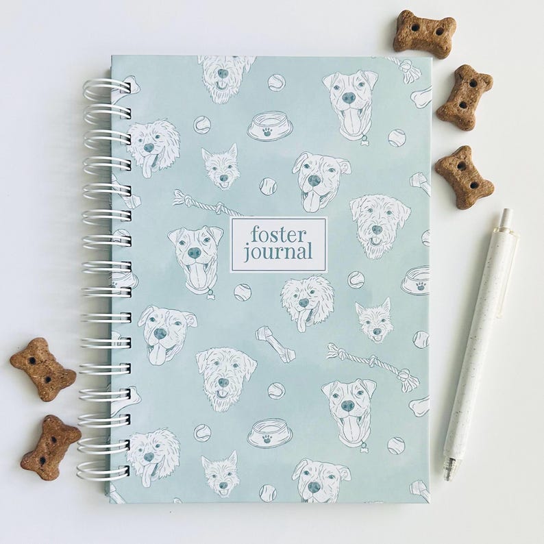 May include: A light blue spiral-bound journal with a repeating pattern of dog faces, tennis balls, and bowls. A central white rectangular label features the text "foster journal" in dark blue. The journal is accompanied by a white pen.