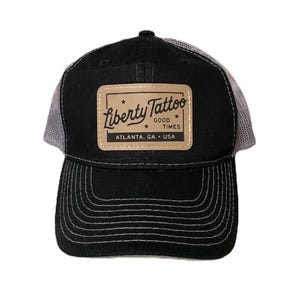 May include: Black and gray trucker hat with a brown leather patch that says "Liberty Tattoo Good Times Atlanta, GA - USA".