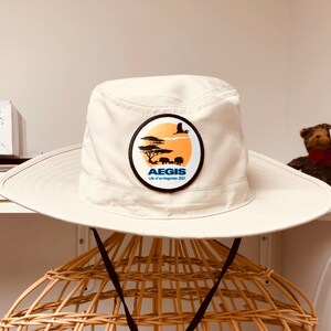May include: A white safari hat with a brown brim and a blue and white logo that says "AEGIS Life of an Aegonian 2021" with a silhouette of a bird and animals in a savanna setting.