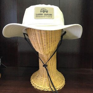 May include: A beige sun hat with a black chin strap and a brown patch on the front that says "LAWN SAFARI PRODUCTIONS" with a small image of a jeep.