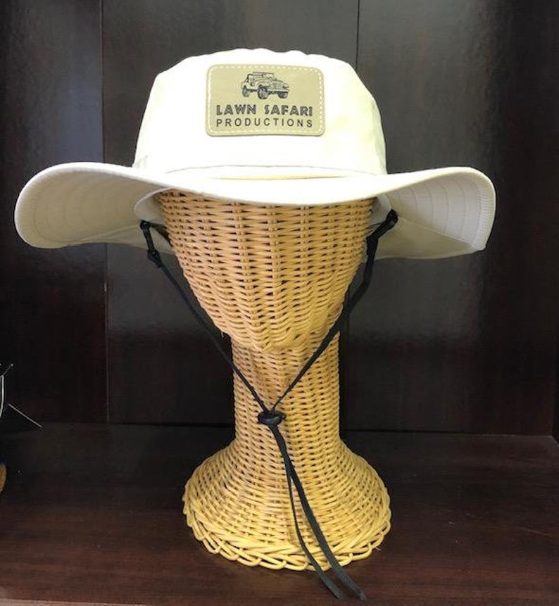 May include: A beige sun hat with a black chin strap and a brown patch on the front that says "LAWN SAFARI PRODUCTIONS" with a small image of a jeep.