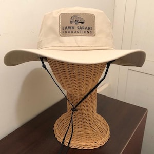 May include: A beige safari hat with a black chin strap and a brown leather patch that says "LAWN SAFARI PRODUCTIONS" with a small image of a jeep.