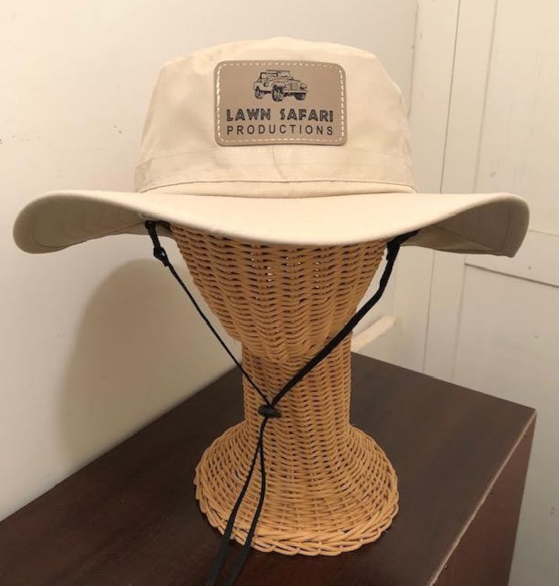 May include: A beige safari hat with a black chin strap and a brown leather patch that says "LAWN SAFARI PRODUCTIONS" with a small image of a jeep.