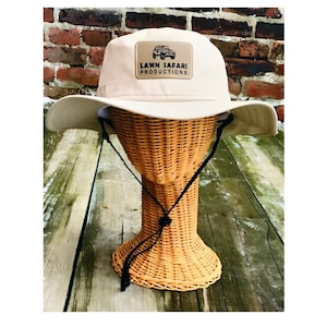 May include: A beige sun hat with a black chin strap and a brown patch with the text "LAWN SAFARI PRODUCTIONS" and a small image of a jeep.