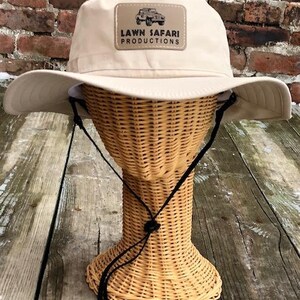 May include: A beige sun hat with a brown patch that says "LAWN SAFARI PRODUCTIONS" and a picture of a jeep.
