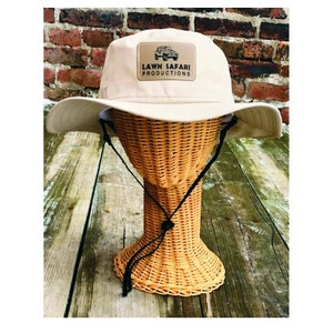 May include: A beige canvas bucket hat with a brown patch on the front that says "LAWN SAFARI PRODUCTIONS" with a small image of a jeep.