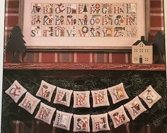 May include: A book cover titled "The Prairie Schooler" featuring a framed cross-stitch alphabet sampler with Christmas-themed designs. Below, a garland of small, embroidered pillows spells out "CHRISTMAS".