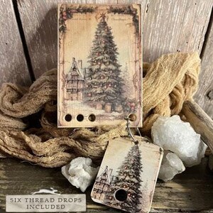 Peut inclure: Une décoration de Noël en bois de style vintage avec une image peinte à la main d'un sapin de Noël et d'une maison. L'ornement comprend six gouttes de fil et fait partie de la collection Smart Thread Keeps. Le texte "OH CHRISTMAS TREE" est imprimé sur l'ornement.