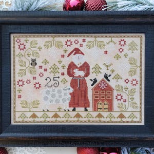 May include: A cross-stitch pattern featuring a red-robed Santa Claus holding a white lamb, a white sheep, a small red house with a white door, and the number 25. The pattern is surrounded by green trees, red stars, and brown branches.