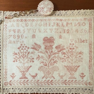 May include: A white linen sampler with a pink floral design and the text "A B C D E F G H I J K L M N O P Q R S T U V W X Y Z 1 2 3 4 5 6 7 8 9 0 + M Marie Wallet"