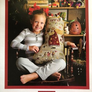 May include: A young girl sits in a rocking chair holding a hand-stitched Christmas stocking with the name "anna" embroidered on it. The stocking is filled with small toys and trinkets. The girl is wearing a striped shirt and grey pants. The stocking is a Shepherd's Bush Printworks design.