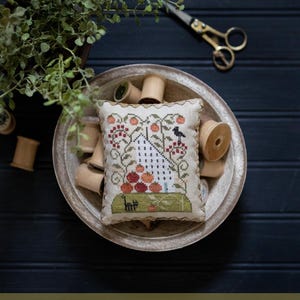 May include: A white cross-stitch sampler with a house, a bird, and a tree with red tomatoes. The sampler is in a wooden bowl with spools of thread. The text "Tomato Hill" is below the image. The text "Plum Street Samplers" is above the image. The text "Original Designs by Paulette Stewart" is below the text "Tomato Hill".