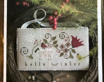 May include: A cross-stitch sampler pattern featuring a white sheep, two red cardinals, and a yellow bird on a branch with red berries and flowers. The text "hello winter" is stitched below the birds. The sampler is framed in a brown wooden frame.