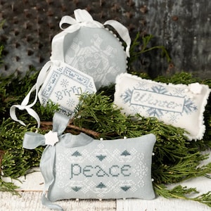 May include: A collection of light blue and white cross-stitched ornaments and pillows with winter-themed designs. The ornaments include a round shape, a tag, and a pillow shape, all with snowflake patterns and the words "birth" and "winter". The pillow in the foreground says "peace".