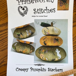 May include: A printed pattern for cross stitch featuring a stack of four pumpkins with Halloween-themed designs. The pumpkins are green, orange, and white with black stitching. The title of the pattern is "Creepy Pumpkin Stackers" and the designer is "Praiseworthy Stitches".