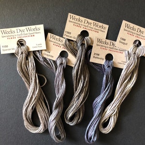 May include: Five spools of Weeks Dye Works embroidery floss in shades of gray and purple. The spools are labeled with the color name and number, including Spanish Moss, Graphite, Pelican Gray, Battleship, and Seagull.