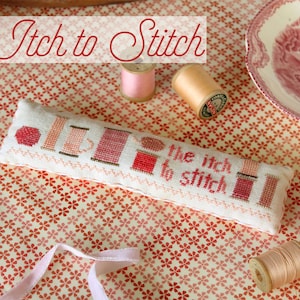 May include: A white needlepoint pillow with the text "The Itch to Stitch" and a design of spools of thread in shades of pink and red. The pillow is on a red and white floral patterned fabric with spools of thread in various colors.