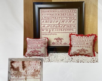 Op de afbeelding: Een vintage kruissteek-sampler met de tekst "M B 1834" en het alfabet in rode draad. De sampler is ingelijst in een donkerhouten lijst en wordt omringd door drie kleinere samplers met hetzelfde ontwerp.