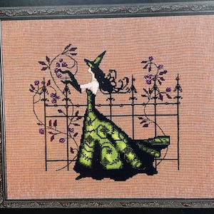 May include: A cross-stitch embroidery of a witch in a green dress and black hat standing in front of a black wrought iron gate with purple berries growing on vines.