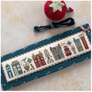 May include: A cross-stitch bookmark featuring a row of colorful houses and floral elements. The bookmark is bordered with a red and white twisted cord. A red strawberry pin cushion, embroidery floss, and scissors are also visible.