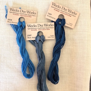 May include: Three skeins of Weeks Dye Works hand-over-dyed cotton floss in shades of blue. The skeins are labeled "2339 Blue Bonnet", "2107 Blue Jean", and "1307 Americana". Each skein is 5 yards long.