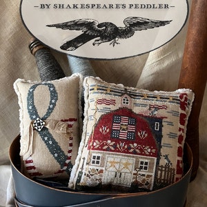 May include: Two rectangular cushions with patriotic designs are displayed in a dark blue container. One cushion has a stitched ribbon and the letter "&", the other a red barn with American flags. The image includes text that reads "& STRIPES" and "-BY SHAKESPEARE'S PEDDLER-".