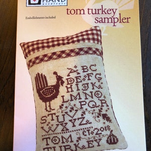 May include: A cross-stitch sampler featuring a turkey with the alphabet, the words "Tom Turkey", and the date "Oct 2018". The sampler is on a cream-colored pillow with a red and white gingham border.