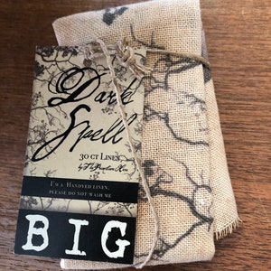 May include: A brown linen tea towel with a black and white floral print. The towel is tied with twine and has a tag that reads "Dark Spell 30 CT Linen by The Preyptare Here I'MA HANDYED LINEN. PLEASE DO NOT WASH ME BIG".