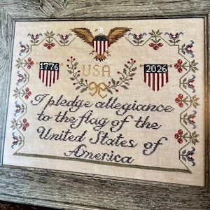 May include: A framed cross-stitch sampler on beige fabric featuring a central bald eagle, the text USA, and the years 1776 and 2026 above the script text I pledge allegiance to the flag of the United States of America, framed by a floral border.