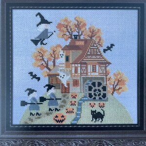 May include: A cross-stitch pattern featuring a spooky house with a black cat, witches, and pumpkins. The house has a brown roof and white walls with windows. The background is a light blue sky with fall leaves.