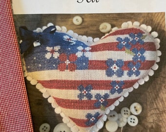 Könnte beinhalten: Ein Kreuzstich-Set für ein patriotisches Herz-Kissen. Das Set enthält roten, weißen und blauen Stoff, Stickgarn und Knöpfe. Das fertige Kissen zeigt ein herzförmiges Design mit einem amerikanischen Flaggenmuster und blauen Blumen. Das Set trägt den Titel "Patriotic Heart Kit" und ist von "Samplers Not Forgotten".