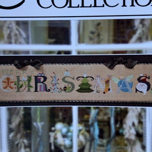 May include: A cross-stitch sampler with the words "Merry Christmas" in colorful embroidery. The letters are decorated with festive imagery, including a gingerbread man, a Christmas tree, and a stocking.