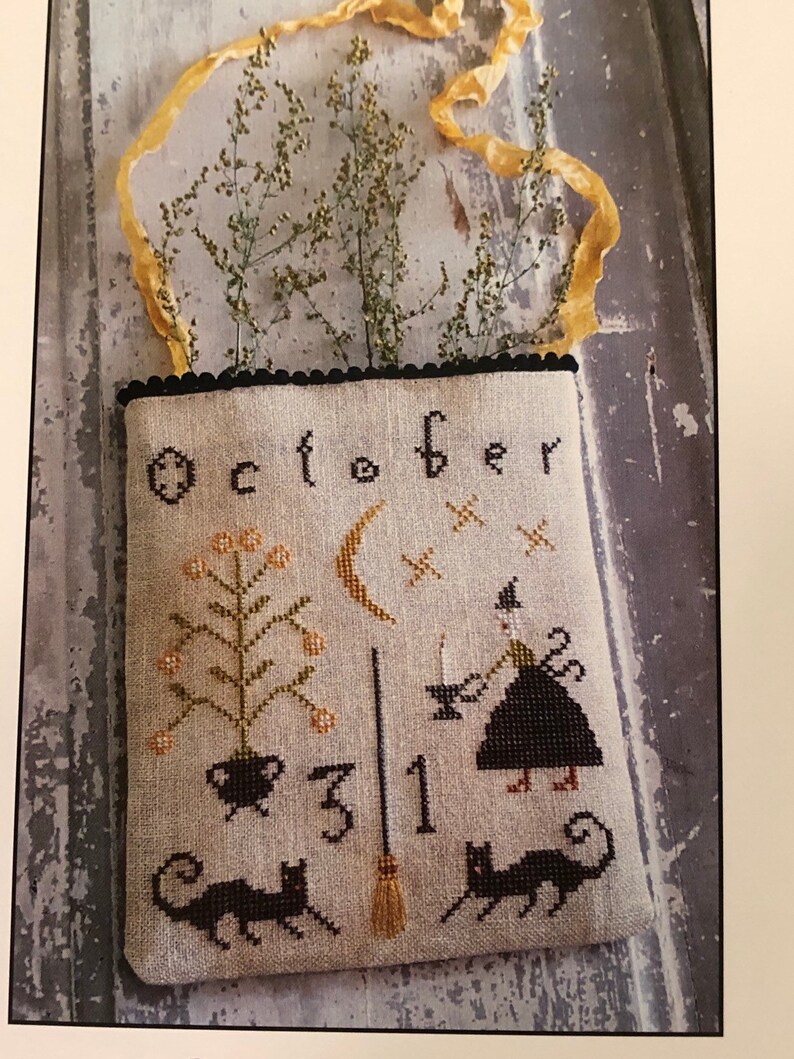 May include: A beige fabric bag with black stitching and a black scalloped border. The bag features an embroidered design of a witch, a cauldron, a broom, a crescent moon, a black cat, and the text "October 31".