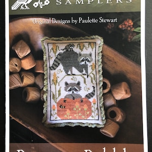 May include: A cross-stitch sampler featuring a group of raccoons on a pumpkin with the text "Raccoon Rabble" and the brand name "Plum Street Samplers".