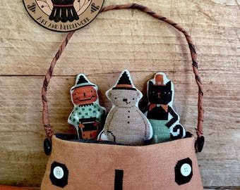 Pode incluir: Uma bolsa de ponto cruz com tema de Halloween com três pequenas figuras recheadas dentro. A bolsa laranja tem um rosto costurado e detalhes de botões. As figuras são uma abóbora, um boneco de neve e um gato preto. O texto "Pumpkin Pals" é visível.