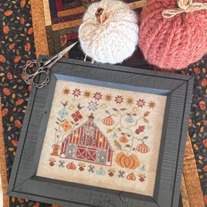 May include: A framed cross-stitch featuring a barn with pumpkins and fall foliage. Two knitted pumpkins, one white and one coral, sit beside the frame. A pair of decorative scissors rests near the frame. The background includes a fall-themed table runner.