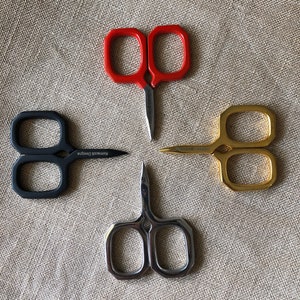 May include: Four pairs of scissors with different coloured handles: red, black, gold, and silver. The scissors are arranged in a cross pattern on a beige fabric background.