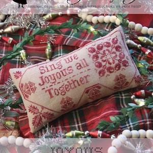 May include: A cross-stitch sampler with the text "Sing we Joyous all Together" on a beige fabric pillow. The pillow is on a red and green plaid fabric with white beads and Christmas ornaments.