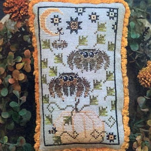 May include: A rectangular, cross-stitched pillow with a Halloween theme. The design features two spiders, a crescent moon, stars, and a pumpkin. The pillow has a light blue background with an orange fringe.