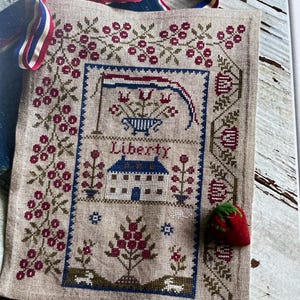 May include: A beige cross-stitch sampler with a patriotic theme, featuring a blue house, floral motifs, and the word "Liberty." A red, white, and blue ribbon is attached to the top. A red strawberry pin is on the right side.
