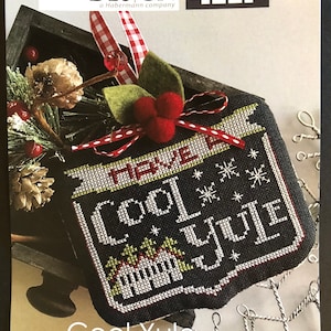 May include: A cross-stitch pattern for a Christmas ornament featuring a black background with white snowflakes and the text "Cool Yule" in white. The ornament is surrounded by a red and white gingham ribbon and a green holly sprig with red berries.