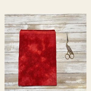 May include: A folded piece of red fabric with a mottled pattern, likely for crafting or sewing. A pair of decorative silver scissors with a bird design rests on a light wood surface.