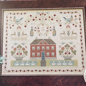 Peut inclure: Une broderie au point de croix encadrée représentant une maison en briques rouges, des compositions florales, des cerfs, des oiseaux et des cygnes. Le motif comprend l'année "1980" et des bordures décoratives. La palette de couleurs comprend le rouge, le bleu, le vert et la crème.