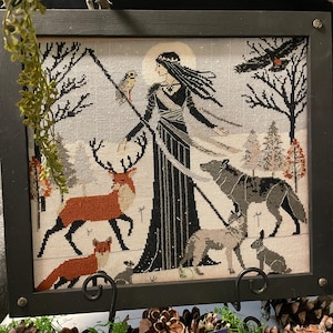 May include: Framed cross-stitch art featuring a woman in a black gown, surrounded by forest animals. The artwork includes a deer, fox, wolf, and rabbit, all rendered in a detailed, folk-art style. The frame is black, and the background is a snowy landscape.